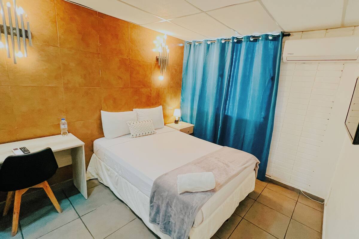 Una habitación de hotel con cama, escritorio, silla, mesita de noche, lámpara y cortina.