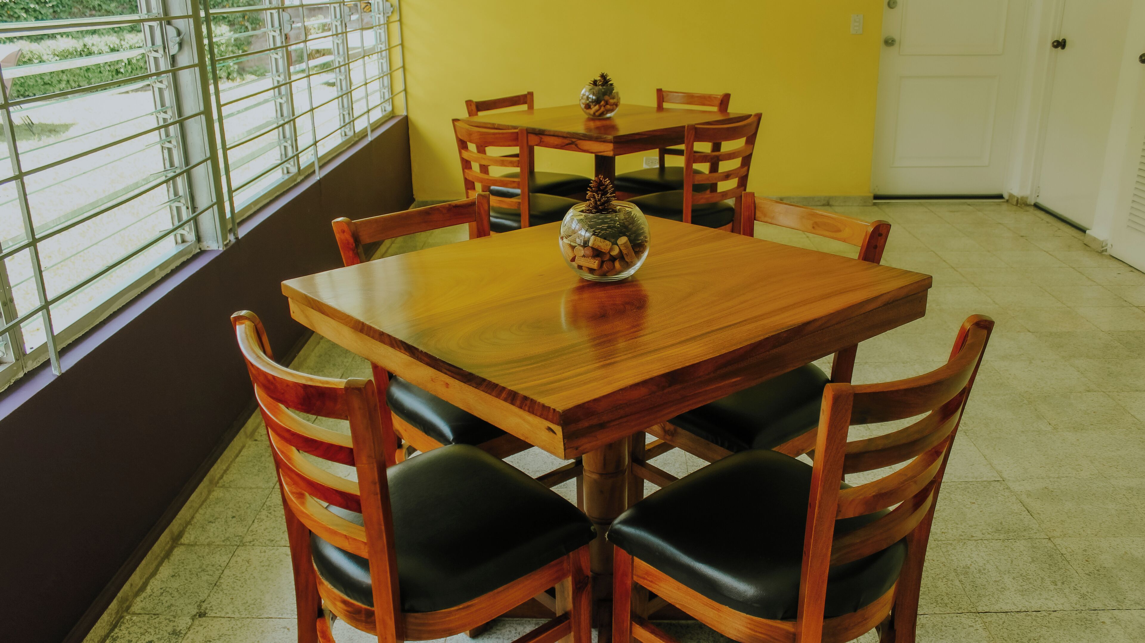 Un comedor con una mesa y sillas de madera, un cuenco de vidrio con piñas, y una vista a través de una ventana con barrotes.