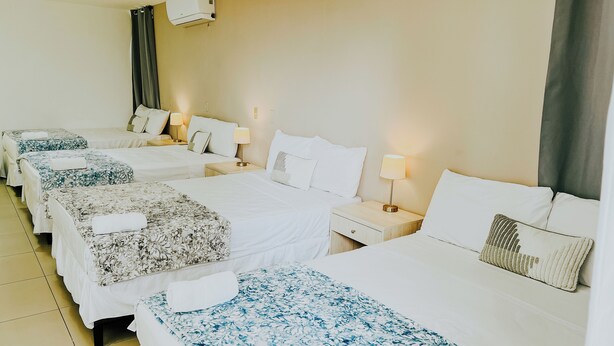 Quarto de hotel com quatro camas de solteiro, cada uma com colcha de padrão floral e travesseiro branco. Há mesas de cabeceira com abajures e ar condicionado fixado na parede.
