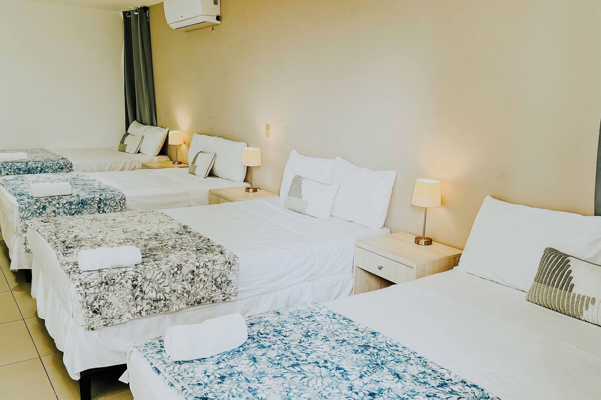 Habitación de hotel con cuatro camas individuales, cada una con colcha de estampado floral y almohada blanca. Hay mesitas de noche con lámparas y un aire acondicionado de pared.