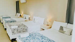 Habitación de hotel con cuatro camas individuales, cada una con colcha de estampado floral y almohada blanca. Hay mesitas de noche con lámparas y un aire acondicionado de pared.