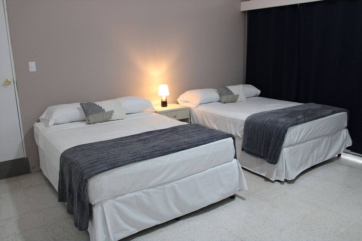 Una habitación de hotel con dos camas individuales, una mesita de noche con lámpara y una cortina oscura en la ventana.