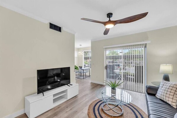 A living room with a flat-screen TV on a white stand, a ceiling fan, a glass coffee table with a plant, a leather sofa, and a view through a sliding glass door to an outdoor area. - The Layover * A Modern living near LV Strip (Las Vegas)