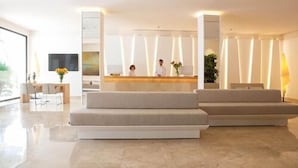 A modern reception area with a reception desk, two staff members, a sofa, and a dining area with chairs and a table.