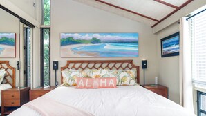 Um quarto com uma cama grande, cabeceira com padrão floral, quadro de uma cena de praia e travesseiro com a palavra