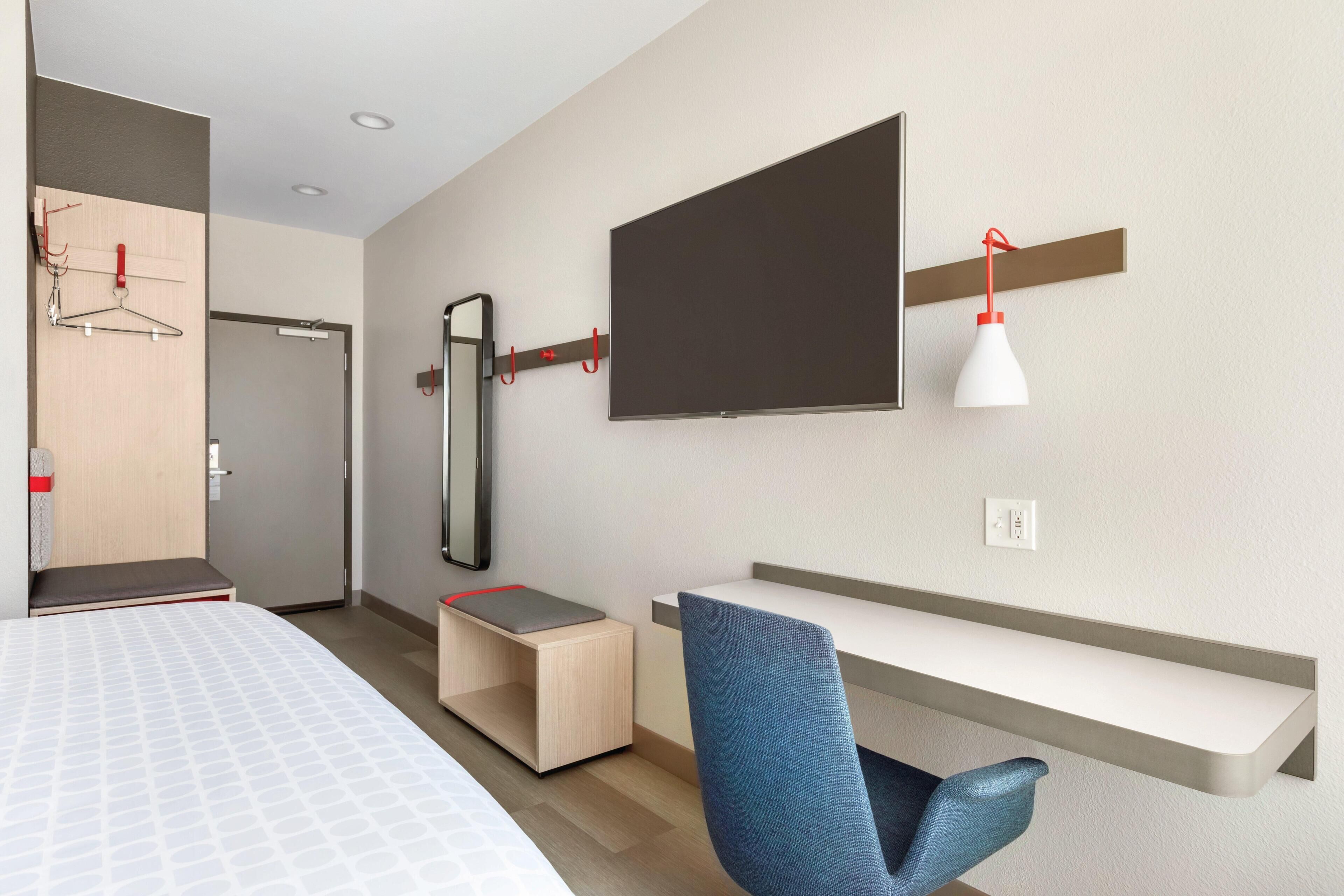 Ein modernes Hotelzimmer mit Bett, Schreibtisch, Stuhl, Fernseher und Spiegel.