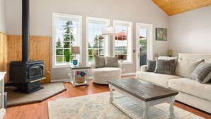 A cozy living room with a wood-burning stove, a sofa, a coffee table, and large windows offering a view of the outdoors. - Ocean Bliss — Coastal Retreat on Skagit Bay (La Conner)