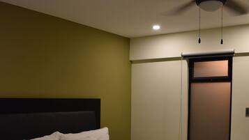 A neatly made bed with white linens in a room with a ceiling fan, a window with blinds, and a plain wall.