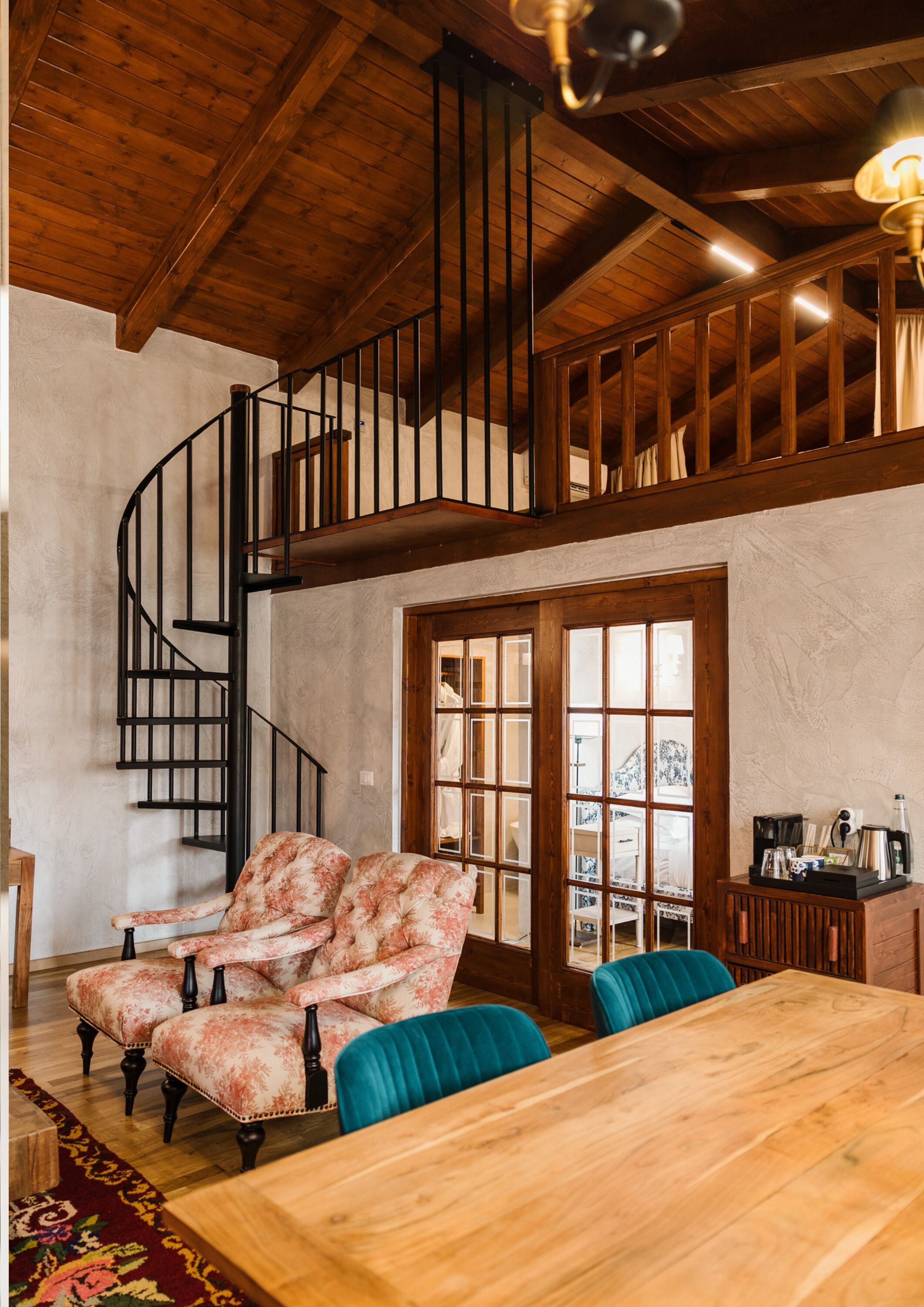 A spacious room with a wooden dining table, two floral armchairs, a blue sofa, and a spiral staircase leading to an upper level.