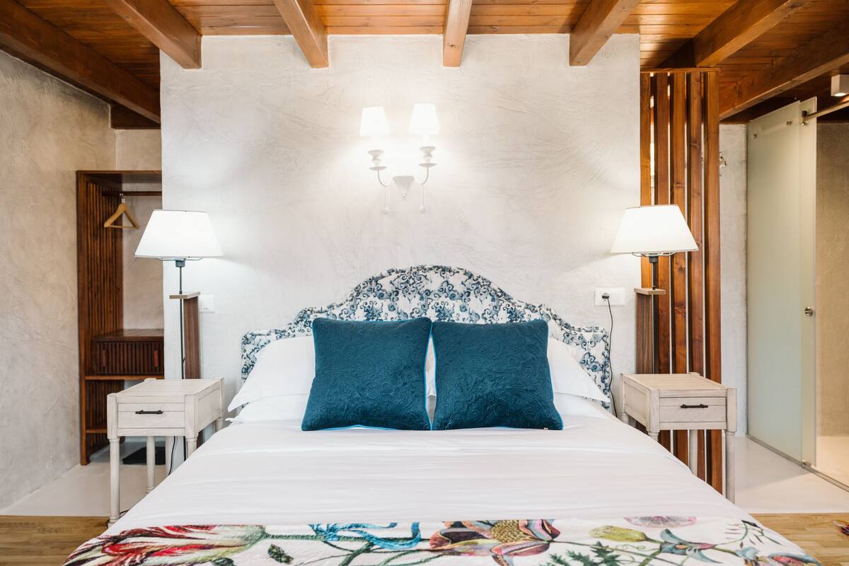 Un dormitorio con techo de madera, una cama con colcha estampada, dos lámparas de noche y una mesita.