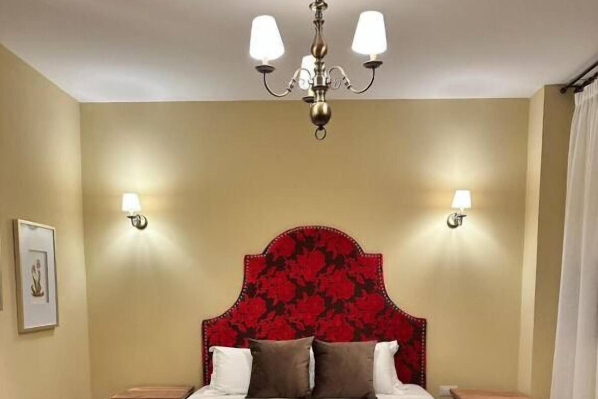 Habitación de hotel con una cama grande, un banco de madera, dos mesitas de noche, un candelabro y lámparas de pared.