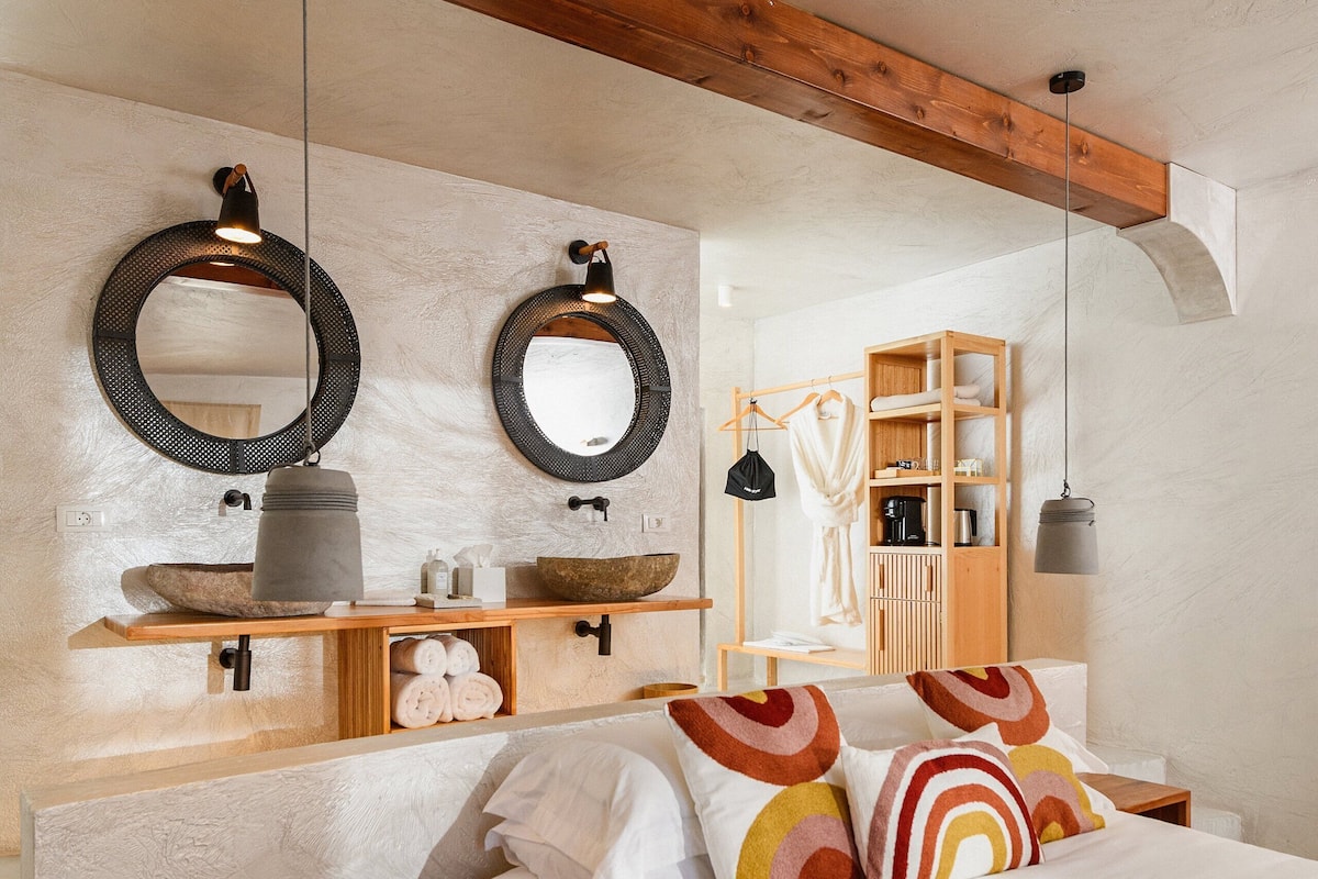 Un dormitorio con cabecera de madera, una mesita de noche, cama con almohadas de colores, una lámpara colgante y un estante de madera con artículos de aseo.