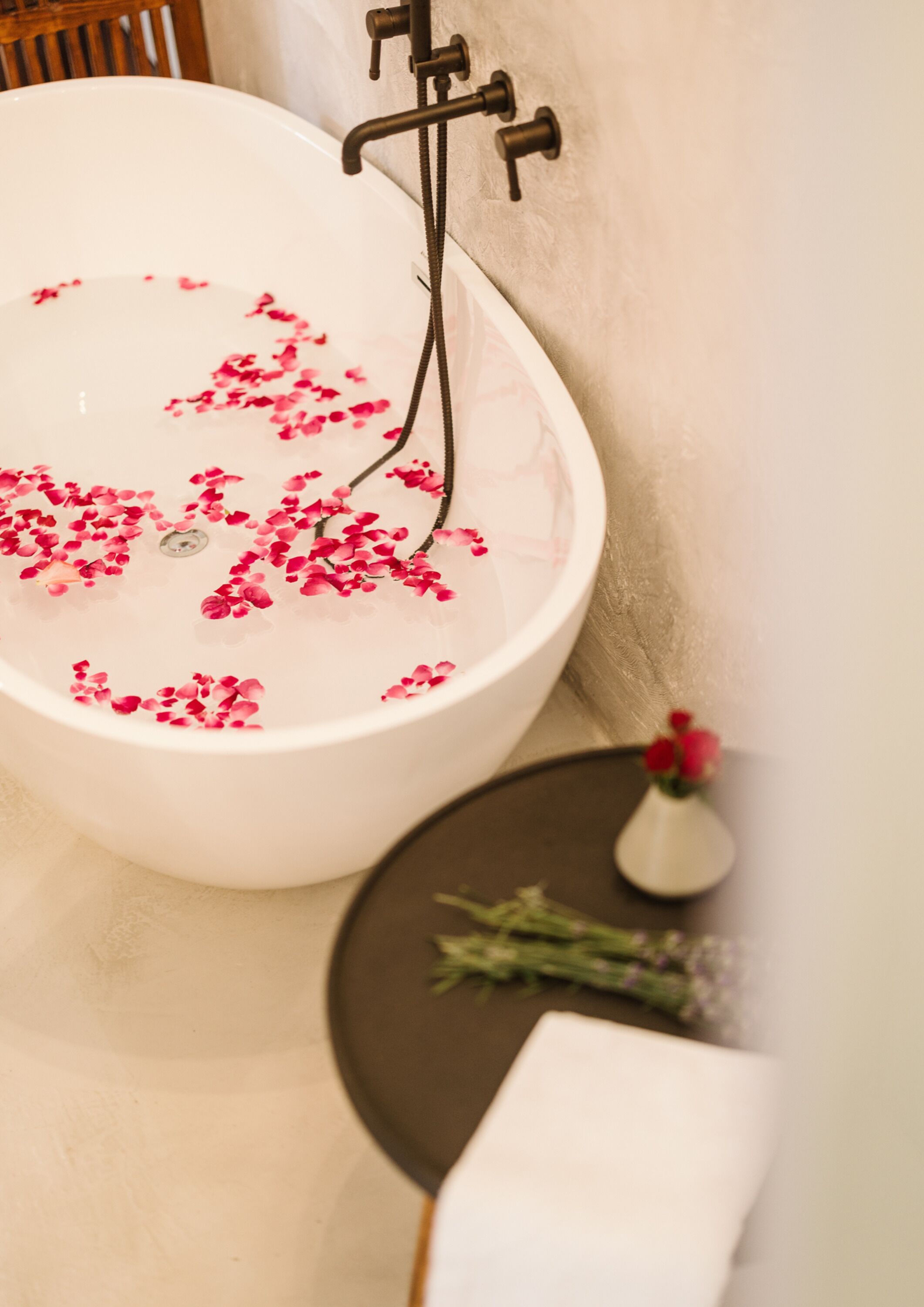 A bathtub with rose petals floating on the water, a small vase with flowers, and a towel on a table.