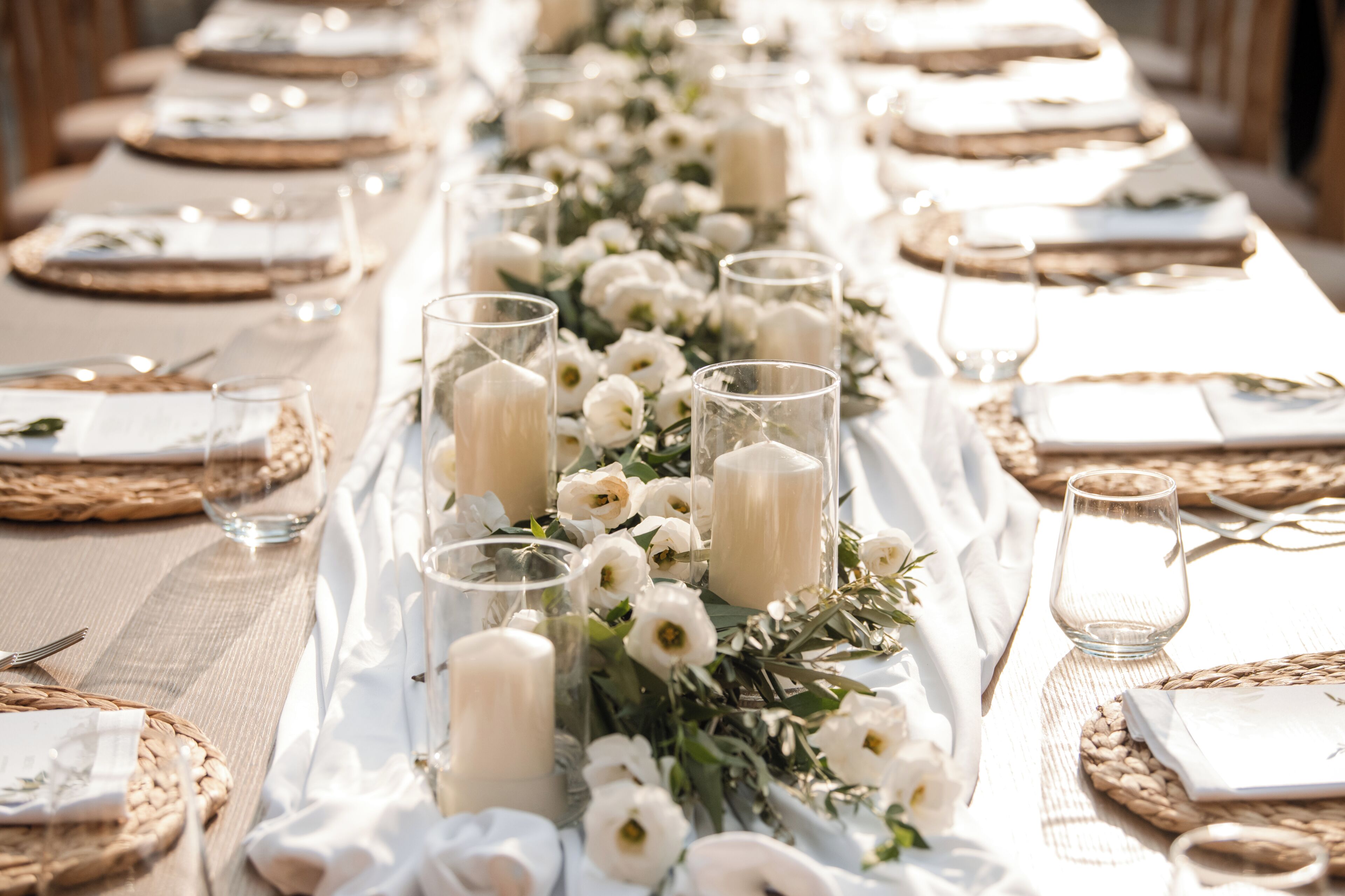 Uma mesa comprida com toalhas brancas, copos transparentes e flores brancas.