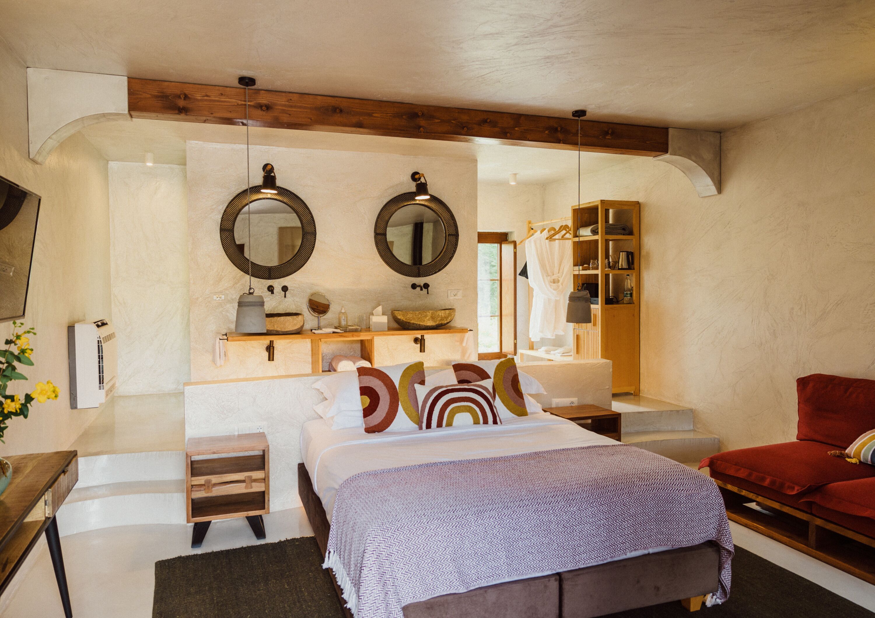 Een slaapkamer met een bed, nachtkastjes, een houten bankje, een rode stoel, een spiegel, een plank met toiletartikelen en een hanglamp.