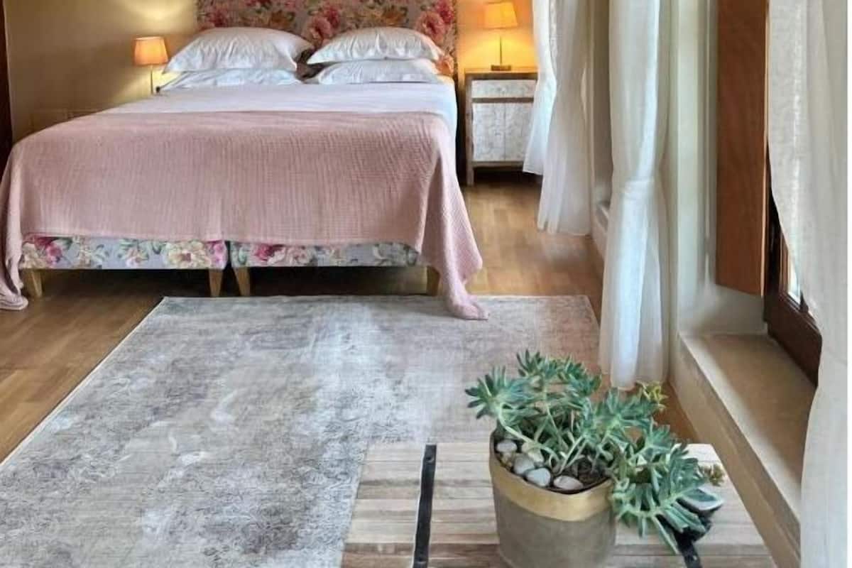 Un dormitorio con cabecera tapizada con motivos florales, una cama con colcha rosa, una mesita de noche con lámpara, una planta en maceta y una ventana con cortinas.