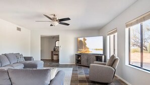 A modern living room with a grey sofa, a grey armchair, and a flat-screen TV on a stand. There is a ceiling fan, a window with a view of the outdoors, and a tiled floor.