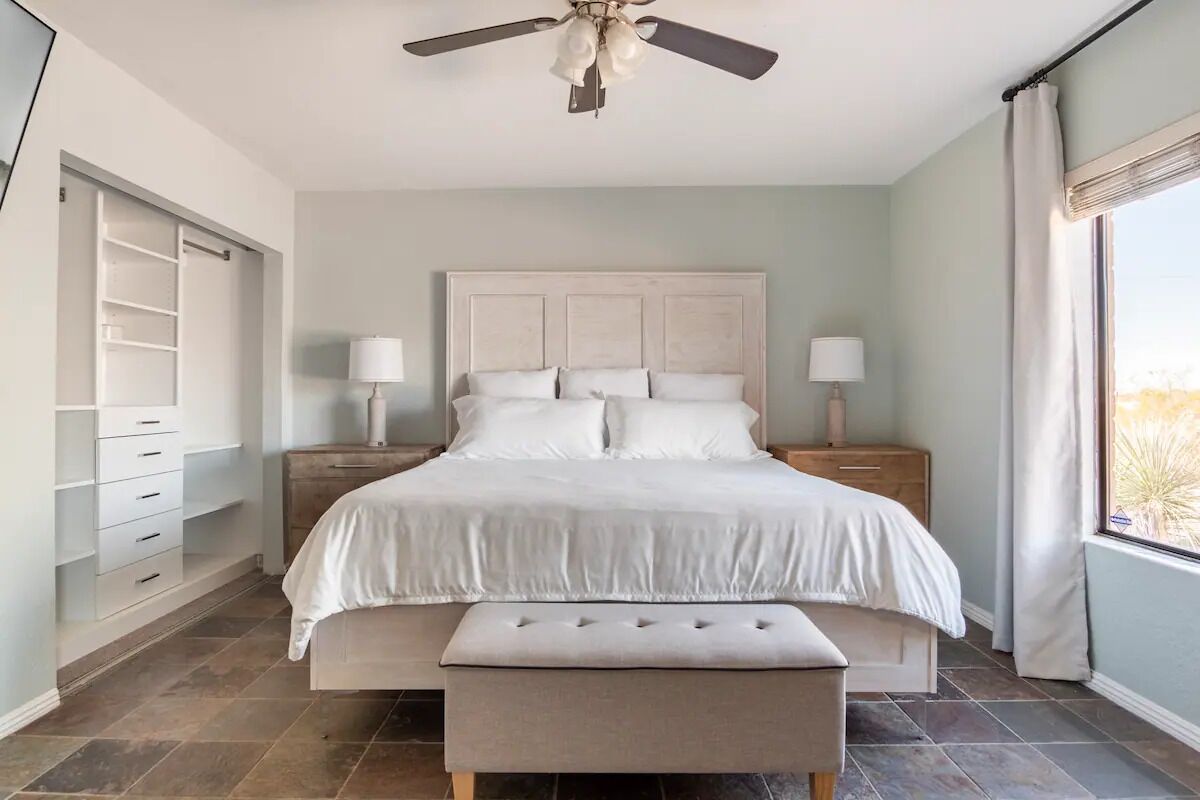 A bedroom with a bed, nightstands, a bench, a ceiling fan, and a window with curtains.