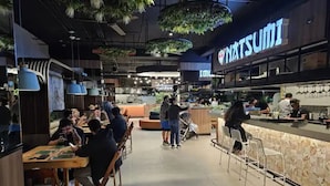 Un interior de restaurante moderno con mesas y sillas, una barra con personal y plantas colgantes.