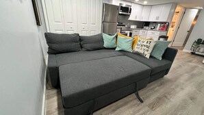 Un salón moderno con un sofá gris, cojines verde azulado y amarillo, y una cocina al fondo.
