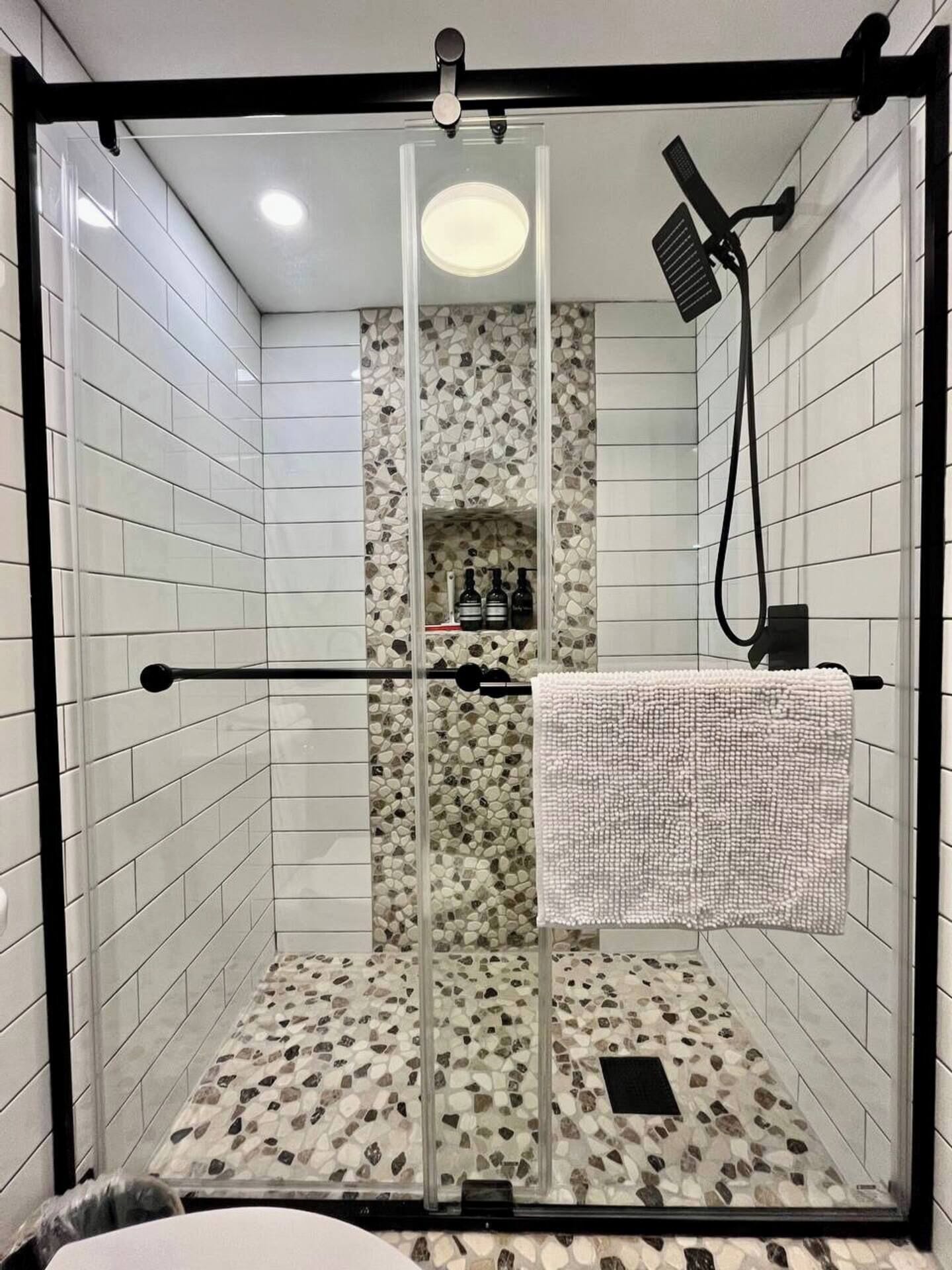 A glass-enclosed shower with a pebble-patterned wall, a towel rack, and a shelf with bottles.