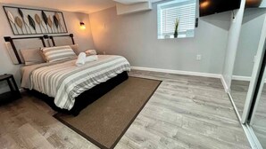 Un dormitorio con cama, mesita de noche, una obra de arte en la pared, ventana con persianas y puerta que da a otra habitación.