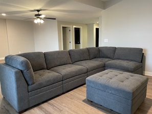 A modern living room with a sectional sofa and an ottoman, wooden flooring, and a ceiling fan.
