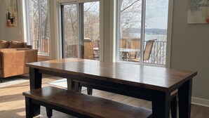 Een ruime kamer met een grote houten tafel en bank, grote ramen met uitzicht op bomen en een waterpartij, en een gezellige zithoek met een bank en stoelen.