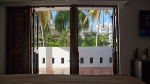Een slaapkamer met een groot bed, een schuifdeur naar een balkon met palmbomen en uitzicht op de stad.