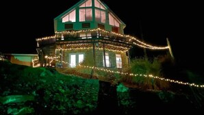 A two-story house with a balcony, surrounded by greenery and illuminated by string lights.