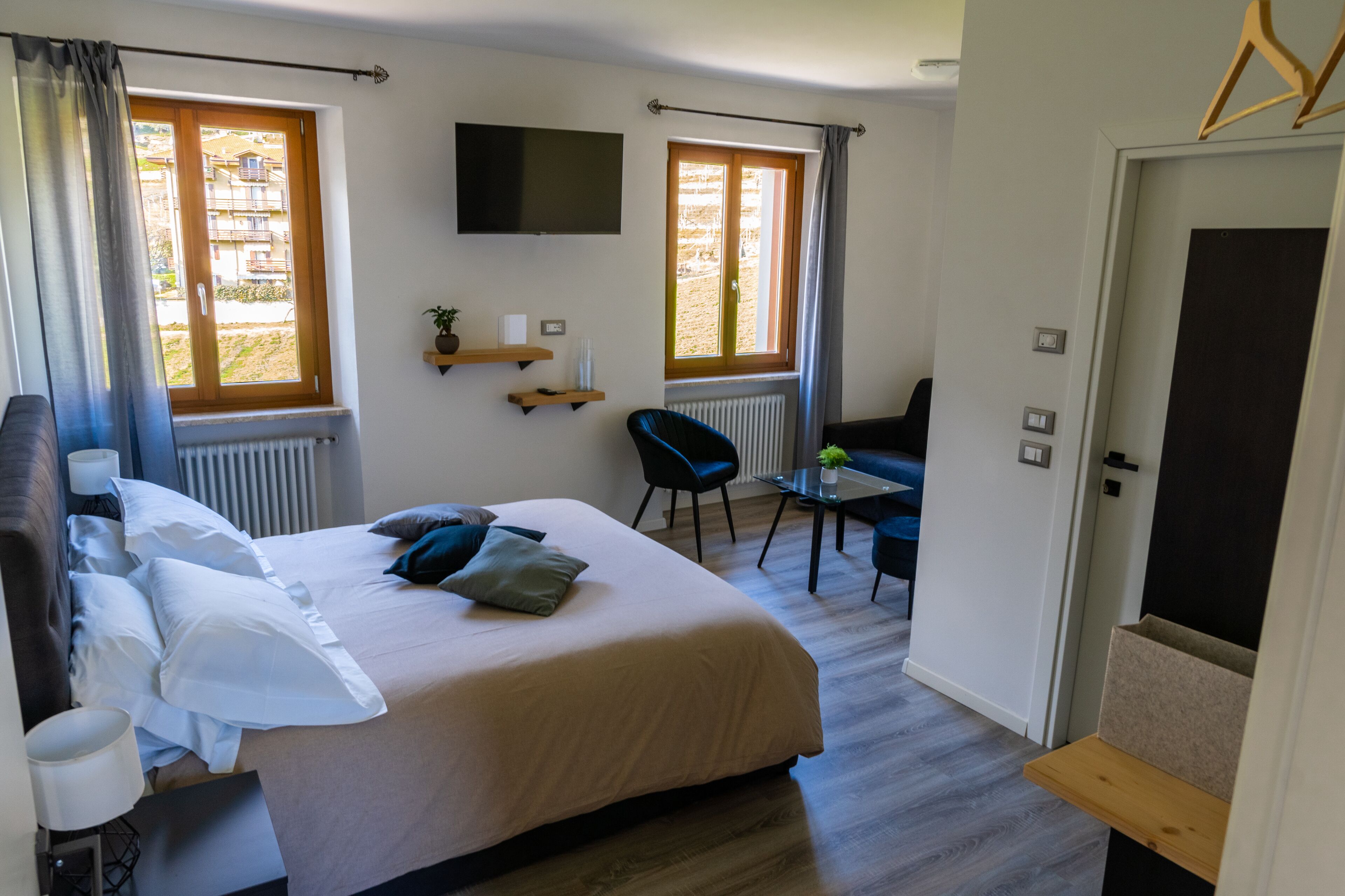 Quarto de hotel com cama, poltrona, mesa, televisão e janelas com cortinas.