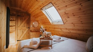A cozy wooden room with a skylight, a bed with neatly folded towels, a tray with pastries, and a small table with a vase of flowers.