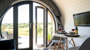 A dining area with a table set for two, a sofa, and a large arched window offering a view of the countryside. - Bedwen -  Glamping pod with Hot Tub sleeping 2 guests (Welshpool)