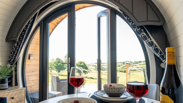 A dining area with a table set for two, two glasses of red wine, a bottle of wine, and a bowl with grapes. - Bedwen -  Glamping pod with Hot Tub sleeping 2 guests (Welshpool)