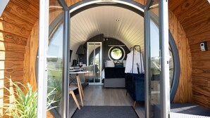 A modern, circular wooden cabin with a glass door entrance, a kitchen area, a dining table, and a cozy seating area. - Bedwen -  Glamping pod with Hot Tub sleeping 2 guests (Welshpool)
