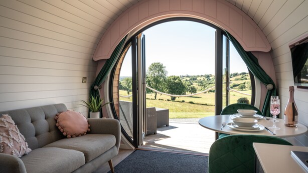 A cozy room with a sofa, a dining table set with plates and a wine bottle, and a view of a green landscape through a large arched window. - Celyn -  Glamping Pod with hot Tub Sleeping 2 People (Welshpool)