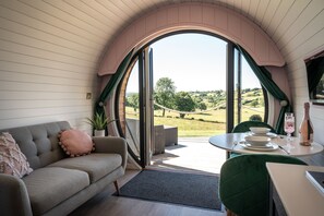 A cozy room with a sofa, a dining table set with plates and a wine bottle, and a view of a green landscape through a large arched window. - Celyn -  Glamping Pod with hot Tub Sleeping 2 People (Welshpool)