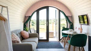 A cozy room with a sofa, a dining table with chairs, and a large arched window offering a view of the outdoors. - Celyn -  Glamping Pod with hot Tub Sleeping 2 People (Welshpool)