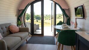 A cozy room with a sofa, a dining table with chairs, and a view of the outdoors through a large arched window. - Celyn -  Glamping Pod with hot Tub Sleeping 2 People (Welshpool)