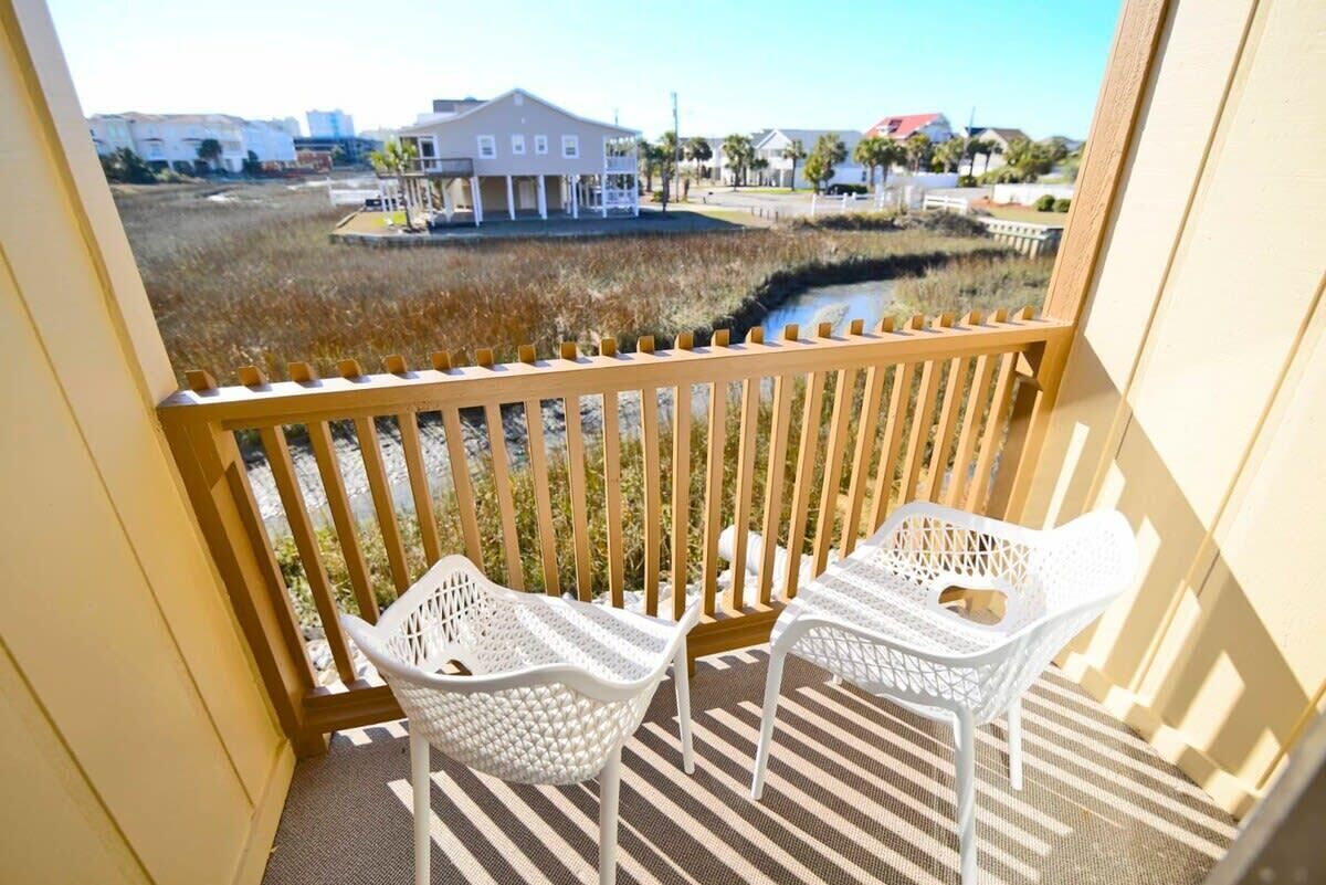 A balcony with two white wicker chairs, a wooden railing, and a view of a building and marshland.