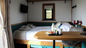 A neatly made bed with a pillow and a folded towel, a bedside table with a plate of food, a bottle, and a mug, and a window with a view of the outdoors. - Cwm Cariad  -  Shepherds Hut with Hot Tub Sleeping 2 guests, Pet Friendly (Knighton)