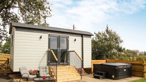A compact, single-story building with a deck, two chairs, a small table, and a hot tub. - Cwm Cariad  -  Shepherds Hut with Hot Tub Sleeping 2 guests, Pet Friendly (Knighton)