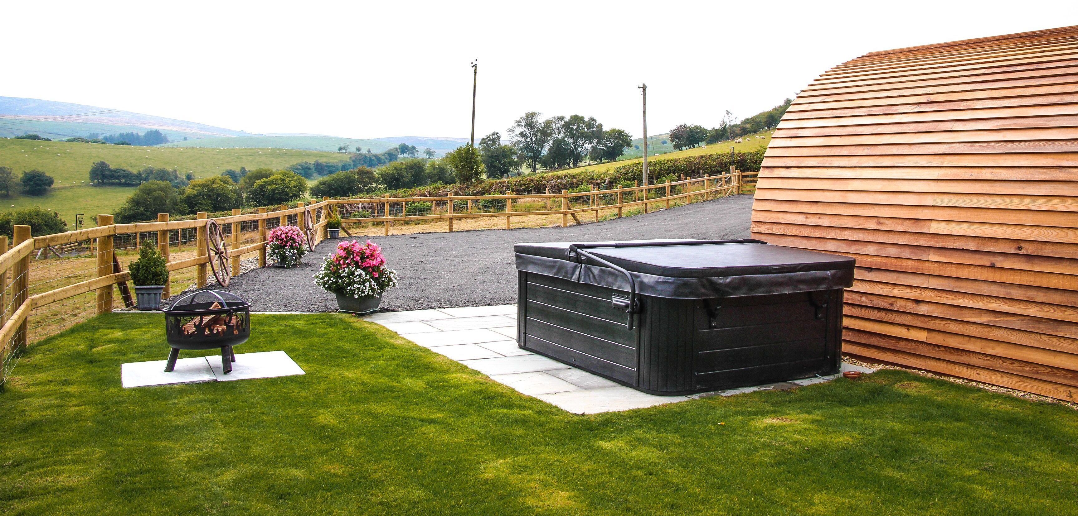 A patio area with a hot tub, a fire pit, and a wooden fence overlooking a green landscape.