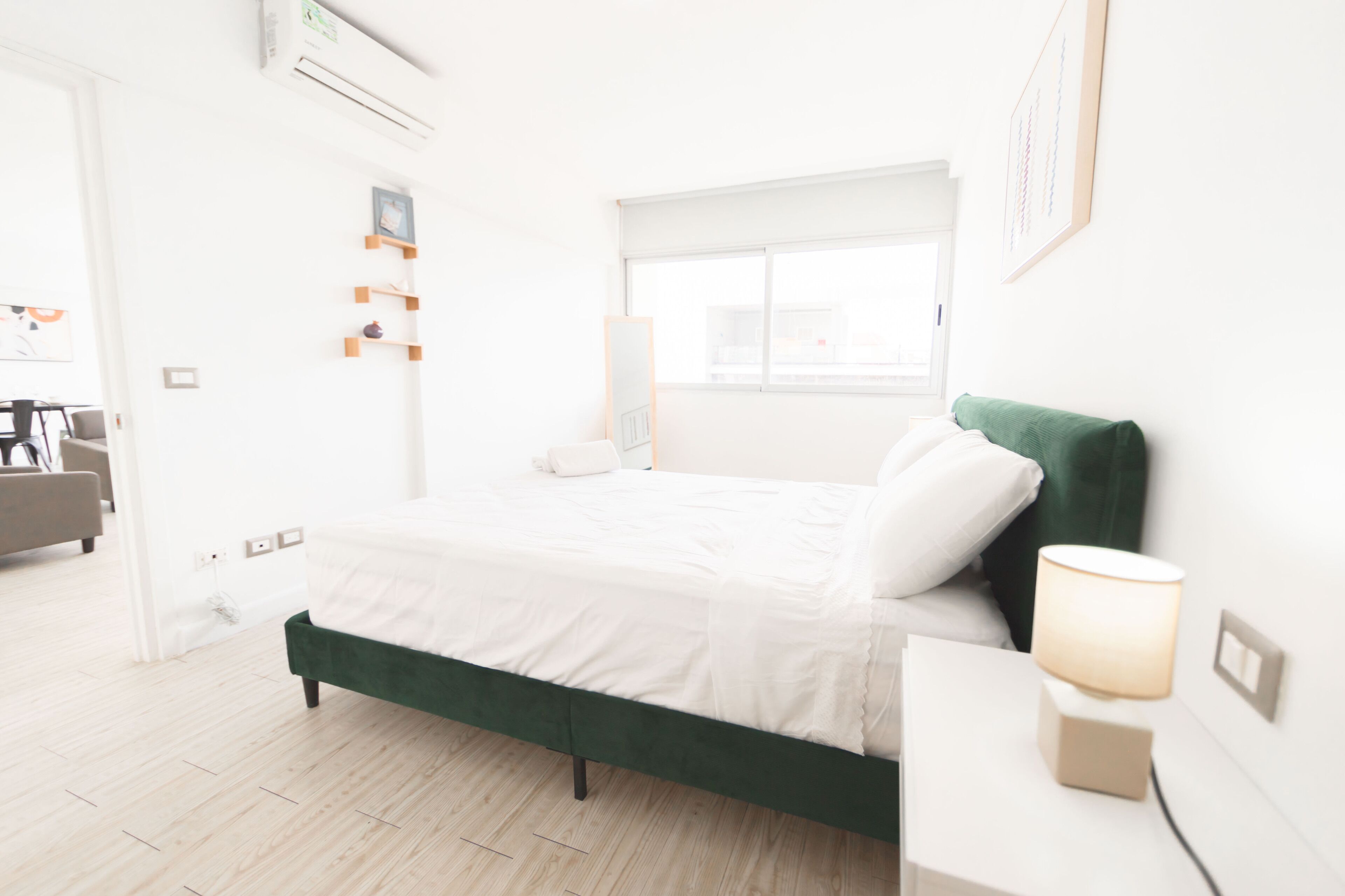 Una habitación de hotel moderna con una cama tapizada en verde, una lámpara de noche y una ventana con vistas.