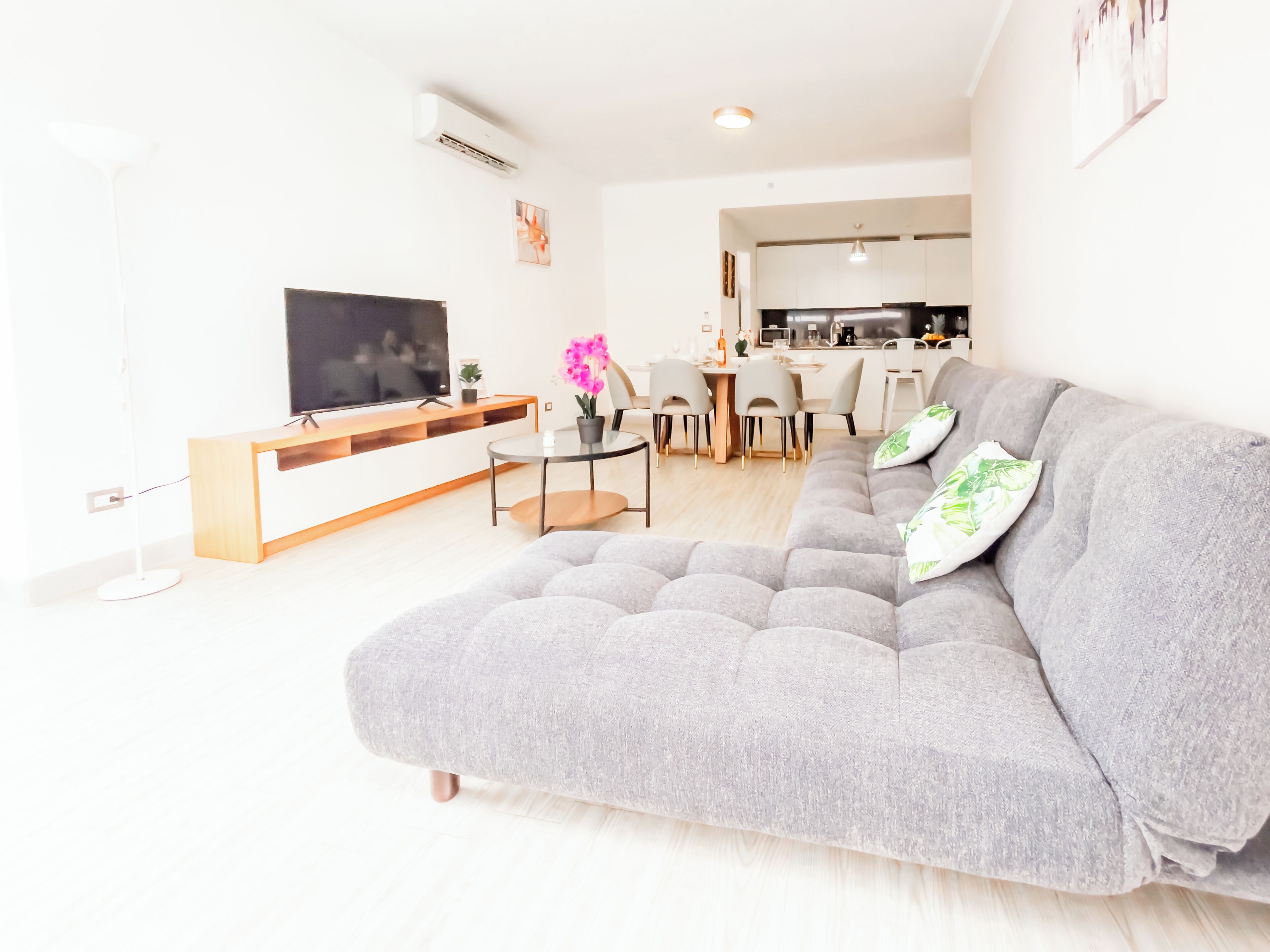 En moderne stue med en grå sofa, et fladskærms-tv, et spiseområde og et køkken i baggrunden.