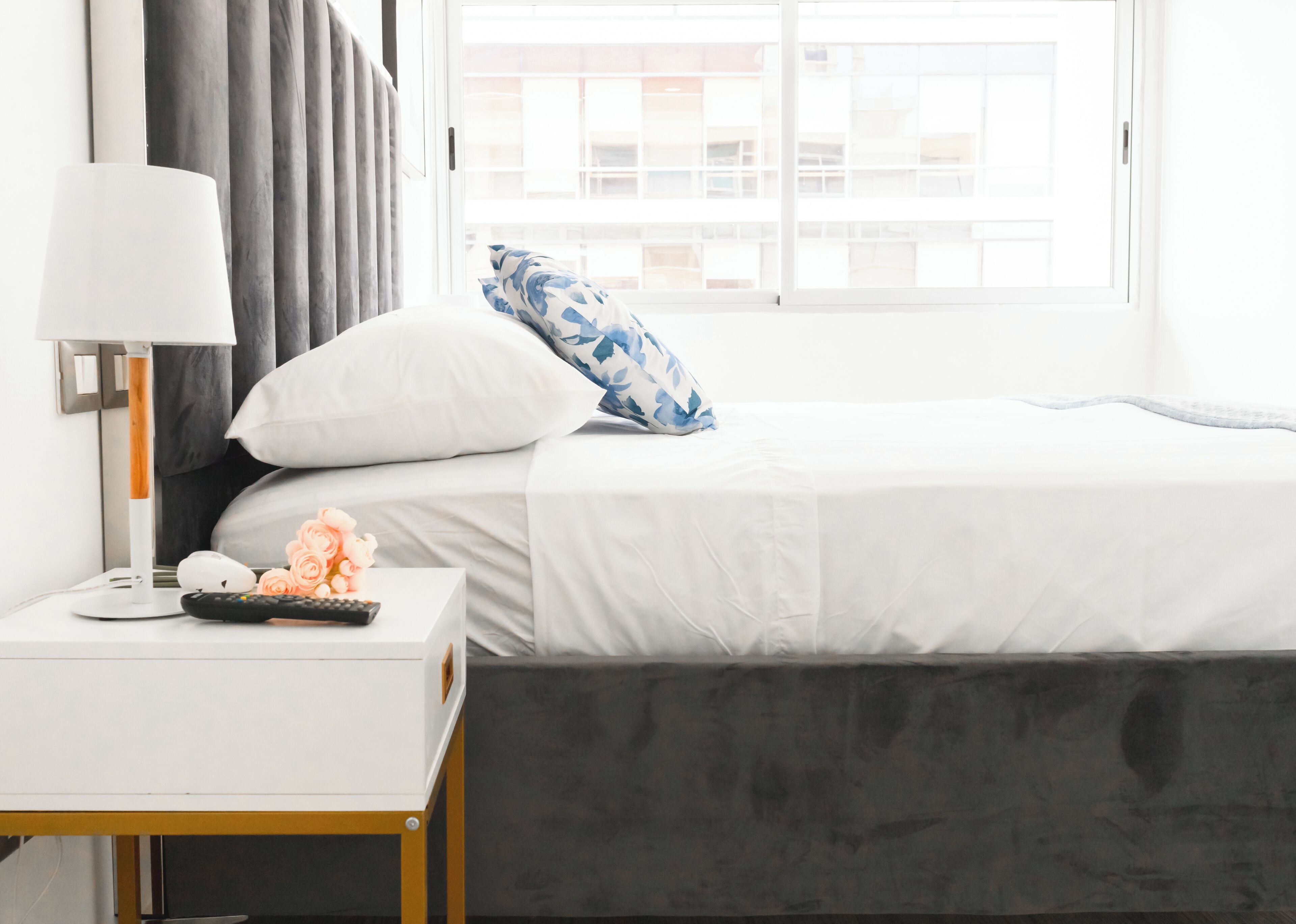 Una cama bien hecha con ropa de cama blanca, cabecera gris y almohada azul y blanca. Se ve una mesita de noche blanca con una lámpara y un control remoto.