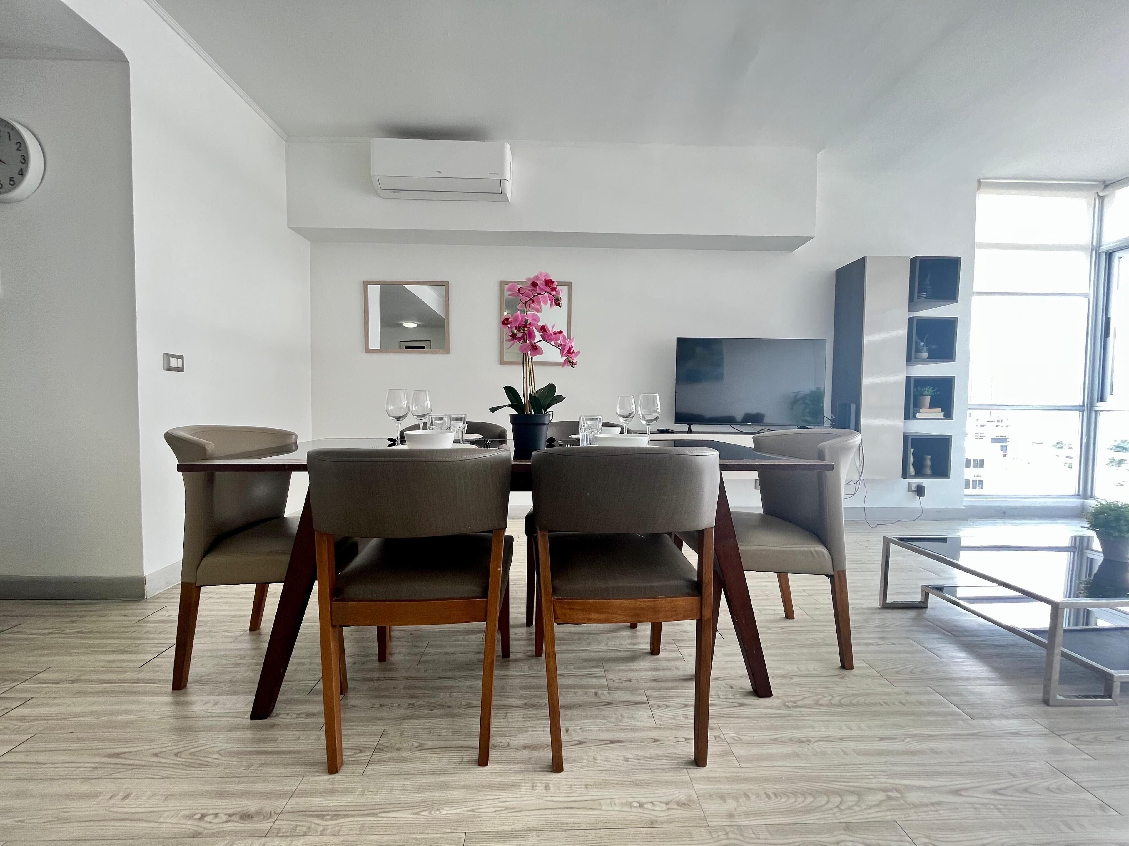 Un moderno comedor con una mesa y sillas de madera, un televisor de pantalla plana y un ventanal con vistas a la ciudad.