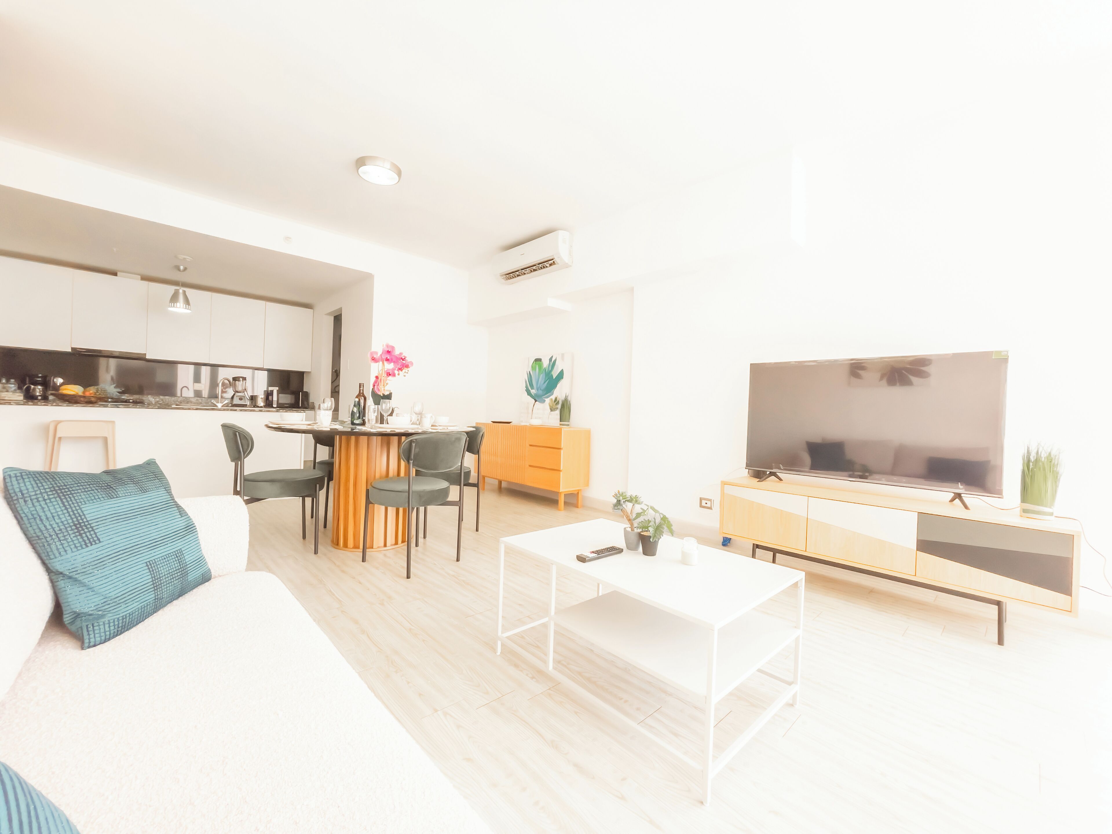 Una sala de estar moderna con un sofá blanco, una mesa de centro y un televisor. Se observa una zona de cocina con armarios y una mesa de comedor.