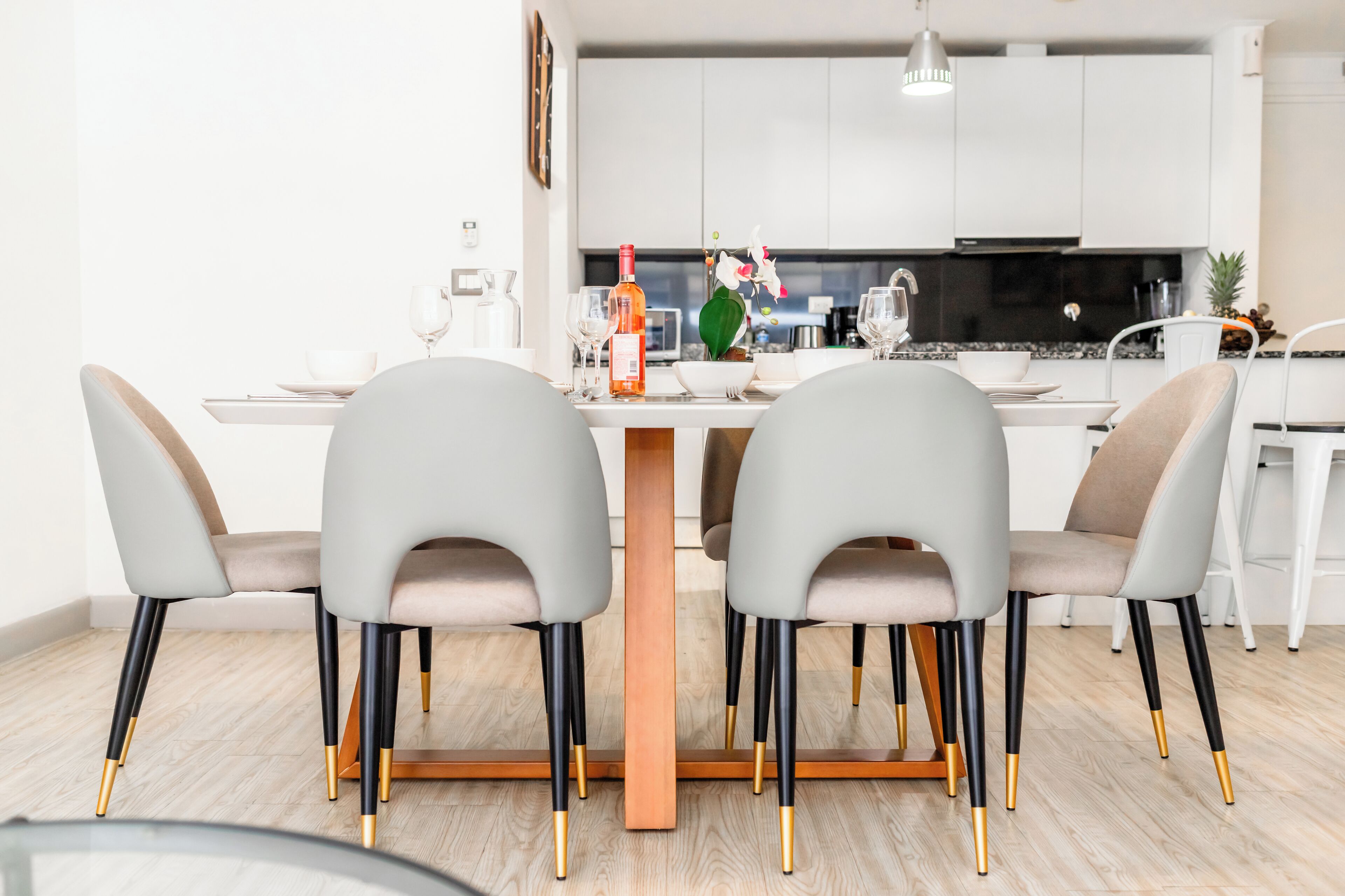 A modern dining area with a wooden table, grey upholstered chairs with gold legs, and a kitchen in the background.