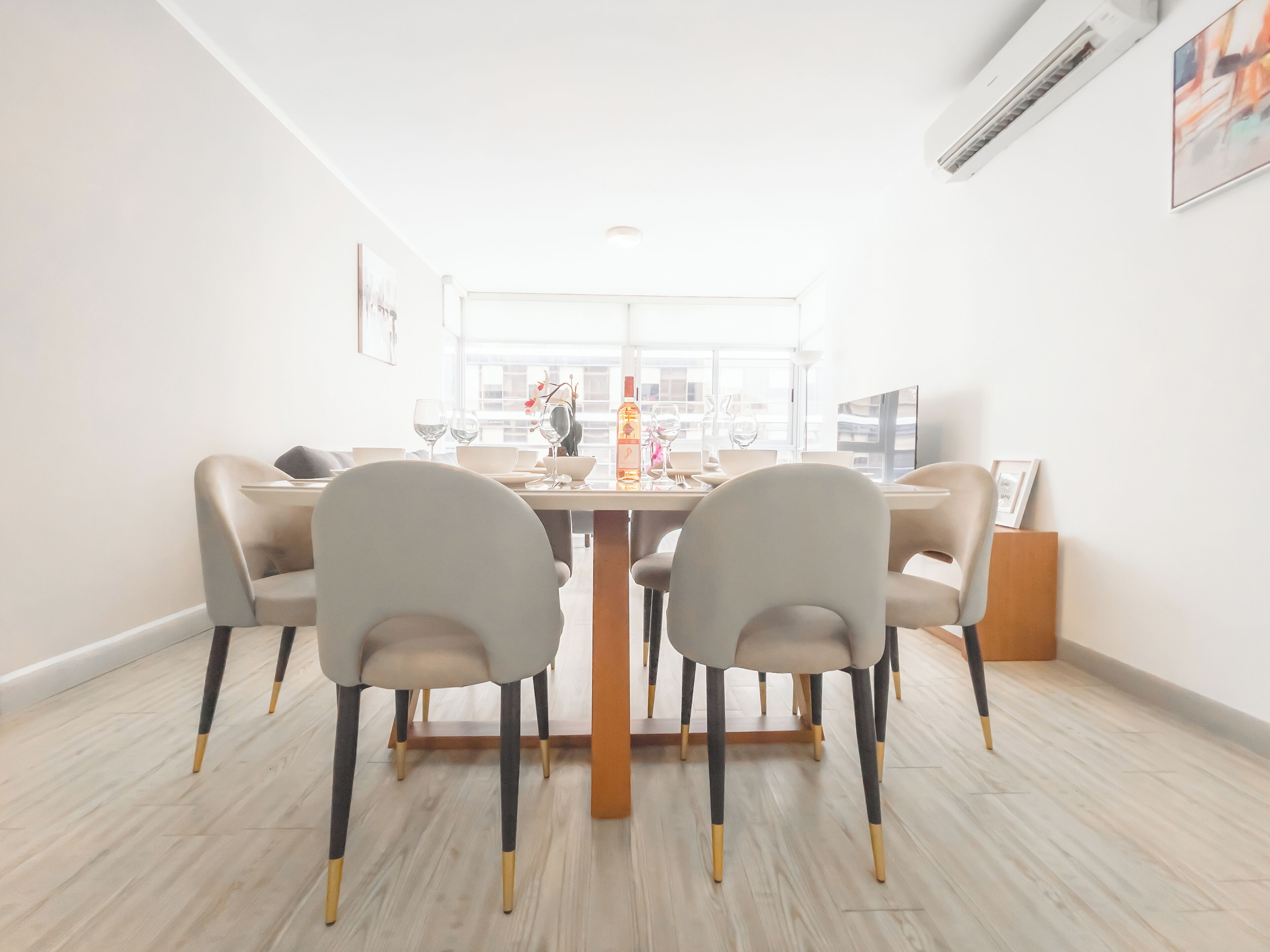 A dining area with a table set for four, surrounded by chairs with gold accents, a window with a view, and a modern air conditioning unit on the wall.